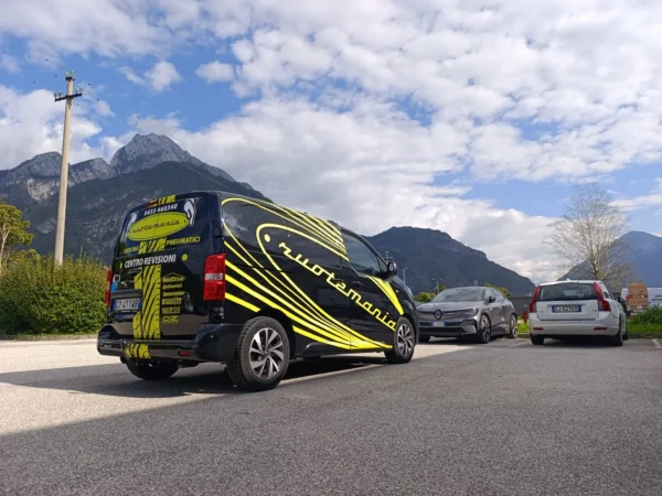 Furgone nero di Ruotemania a Tolmezzo con la grafica gialla a battistrada, visto da dietro con le montagne