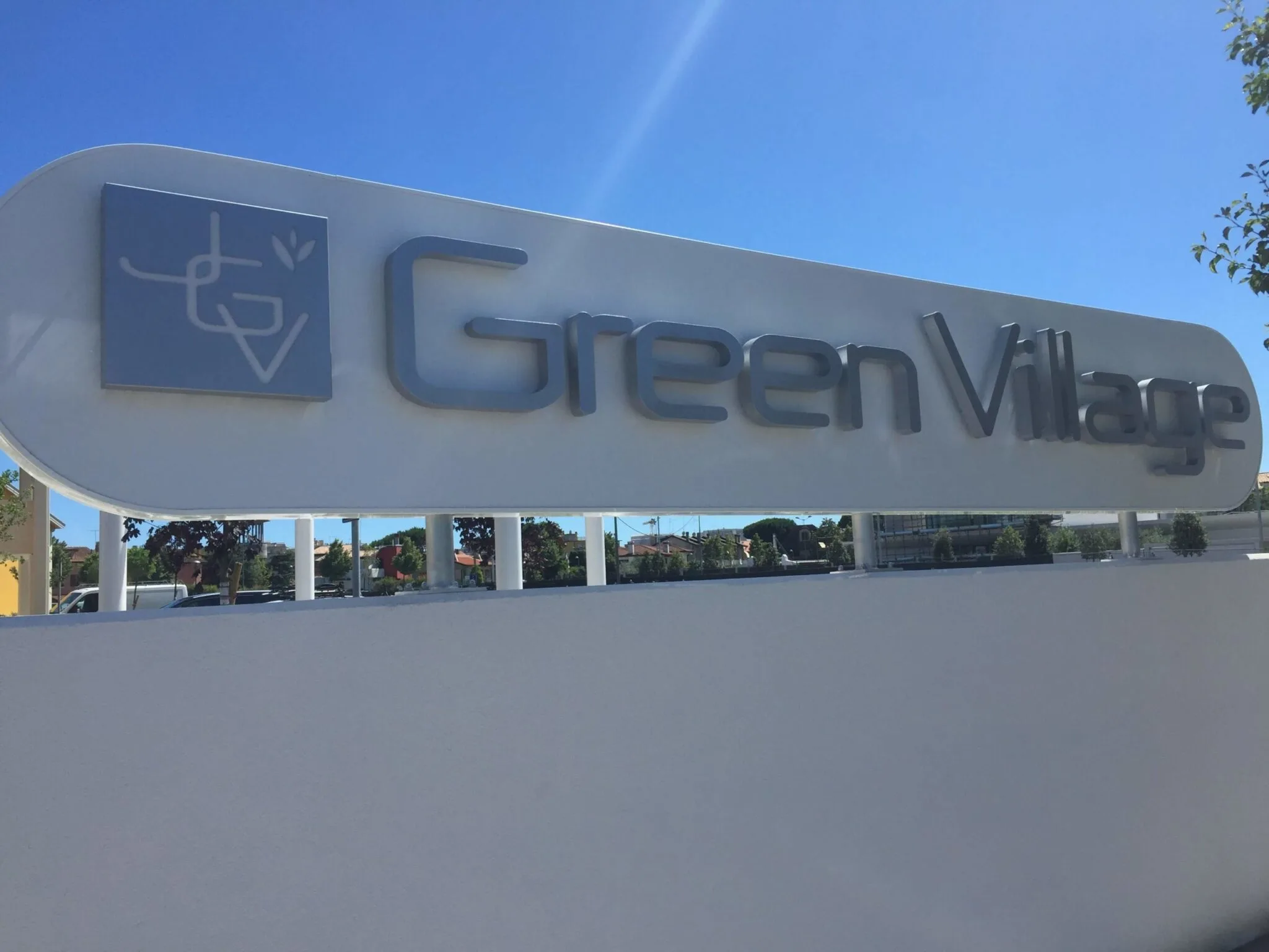 Insegna Green Village in acciaio inox satinato su pannello bianco arrotondato, vista da vicino al sole