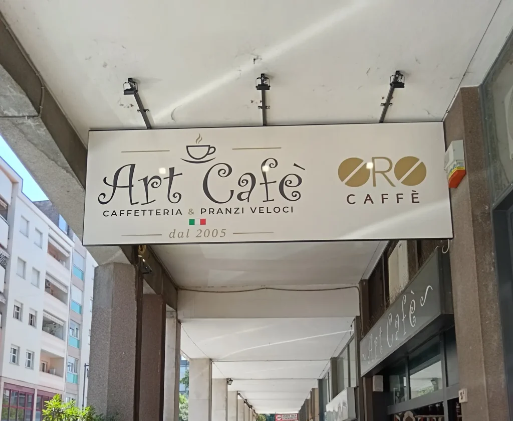 Insegna a pannello di Art Caf&egrave;, caffetteria e pranzi veloci, appesa sotto il portico a Pordenone, col logo Oro Caff&egrave; e i faretti LED neri