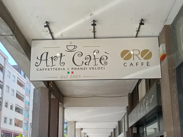 Insegna a pannello di Art Caf&egrave;, caffetteria e pranzi veloci, appesa sotto il portico a Pordenone, col logo Oro Caff&egrave; e i faretti LED neri