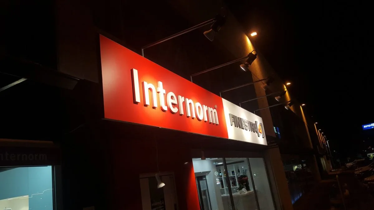 Pannello rosso Internorm con lettere in rilievo e faretti a sbraccio, ripresa serale dalla strada a Udine