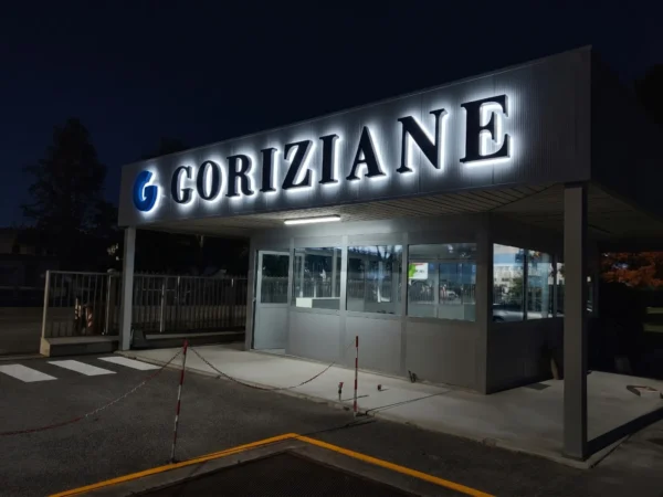 Insegna Goriziane a Villesse di notte: alone bianco a luce riflessa dietro le lettere nere e logo G illuminato di blu