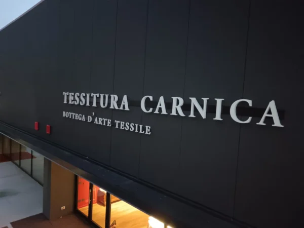 Insegna a lettere singole non luminose "Tessitura Carnica, bottega d'arte tessile" su facciata scura