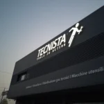 Insegna luminosa Tecnista a lettere singole con l'omino e Tools in action, accesa sulla facciata scura al tramonto, San Giorgio di Nogaro