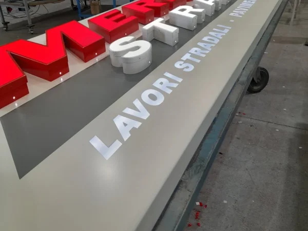 Insegna Merluzzi Strade in costruzione in officina, lettere scatolate rosse e scritta Lavori stradali traforata