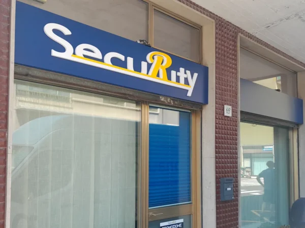 Insegna di Security sopra l'ingresso a Monfalcone: fondo blu, scritta bianca in rilievo con la R gialla rialzata