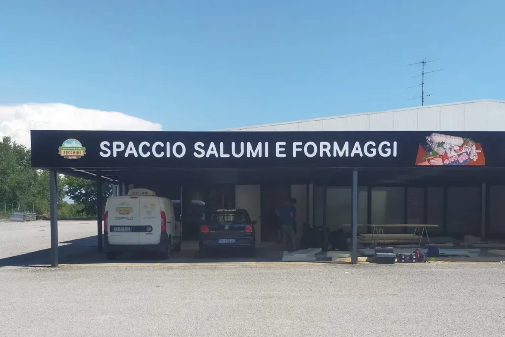 Fascia nera &laquo;Spaccio salumi e formaggi&raquo; lungo la tettoia del Salumificio Zecchini a Spilimbergo, con le foto dei salumi