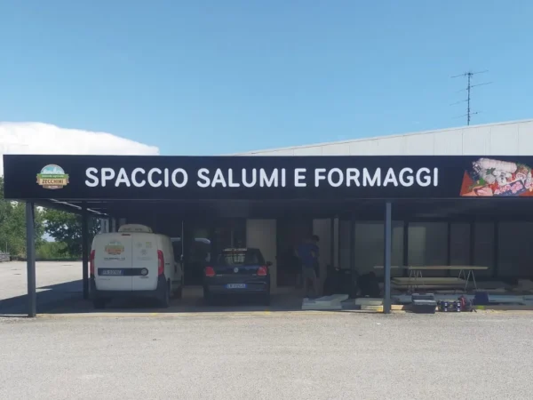 Fascia nera &laquo;Spaccio salumi e formaggi&raquo; lungo la tettoia del Salumificio Zecchini a Spilimbergo, con le foto dei salumi