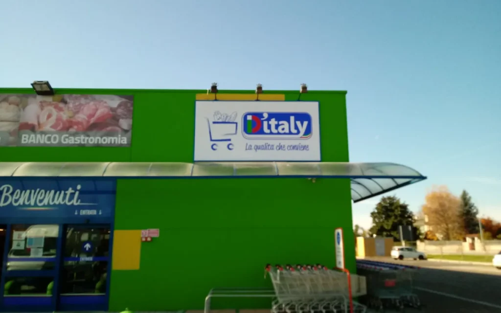 Pannello sagomato D'Italy col carrello sulla facciata verde del supermercato di Fogliano Redipuglia, con i faretti a braccio sopra