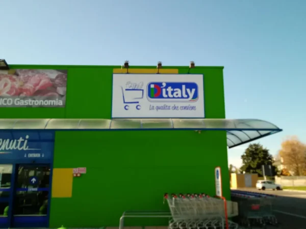 Pannello sagomato D'Italy col carrello sulla facciata verde del supermercato di Fogliano Redipuglia, con i faretti a braccio sopra