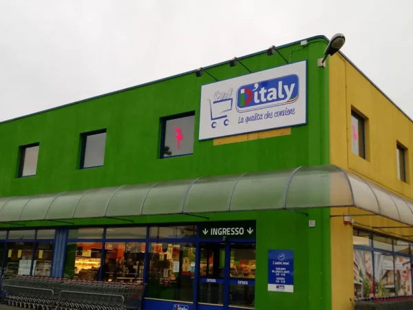 Pannello sagomato D'Italy col carrello sull'angolo della facciata verde del supermercato a Monfalcone, con i faretti a braccio