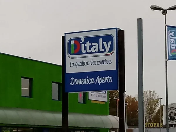 Il cassonetto luminoso D'Italy «Domenica aperto» sul palo, verso il parcheggio
