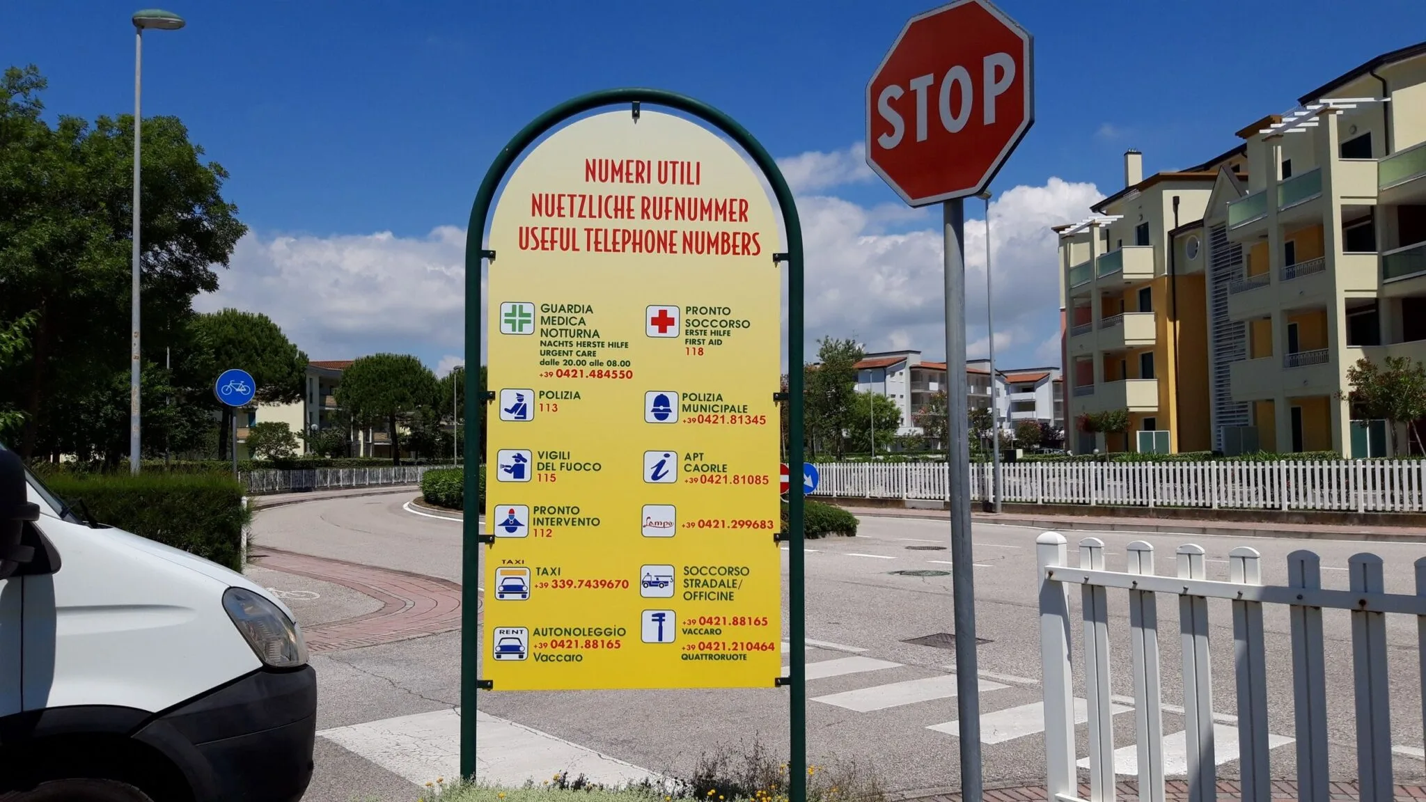 Cartello Numeri utili in tre lingue con simboli per soccorso e polizia, lungo la strada del villaggio